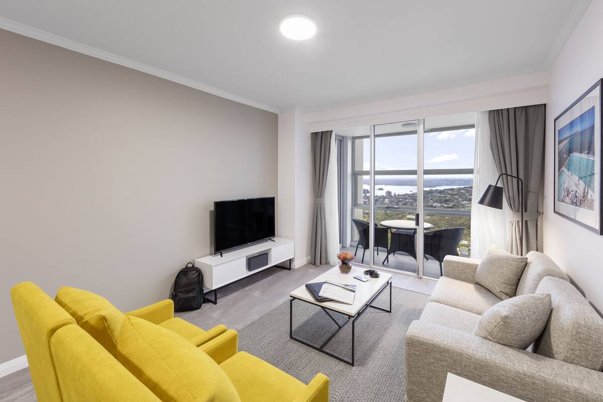 Un salon moderne avec un canapé, des fauteuils jaunes, une table basse, un téléviseur à écran plat et un balcon offrant une vue sur la ville.
