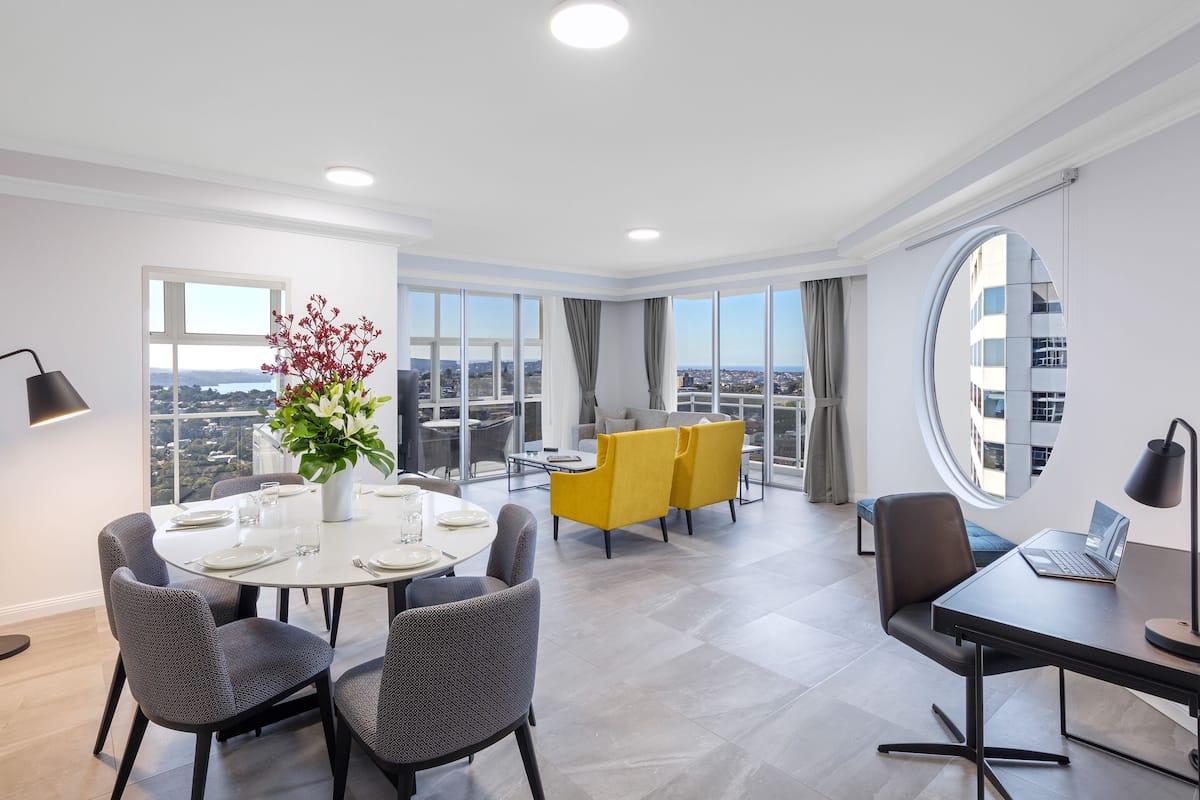 Une chambre d’hôtel moderne avec un coin repas, un ordinateur portable posé sur un bureau et une grande fenêtre donnant sur la ville.