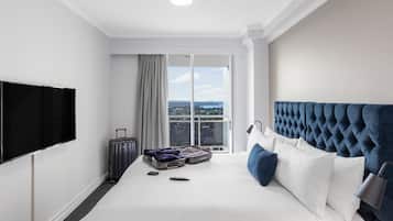 A hotel room with a large bed, a flat-screen TV, a suitcase, and a view of the city through the window.