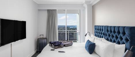 A hotel room with a large bed, a flat-screen TV, a suitcase, and a view of the city through the window.