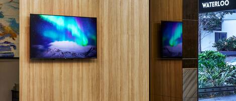 Un vestíbulo moderno con paneles de madera, dos televisores mostrando la aurora boreal, una mesa redonda con sillas y vistas del edificio Waterloo.