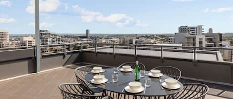 A rooftop terrace with a table set for dining, surrounded by chairs, and overlooking a cityscape.