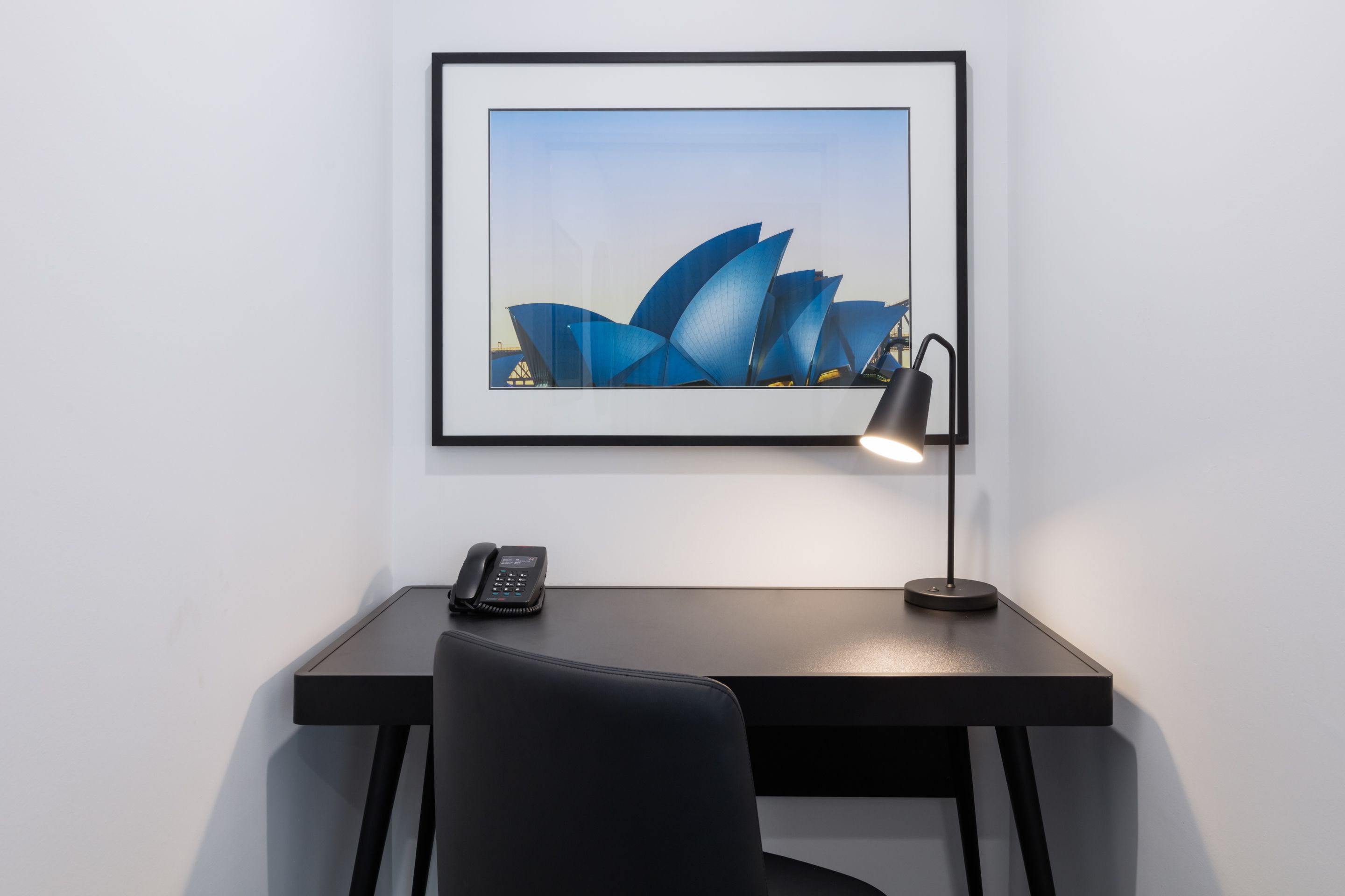 Ein Schreibtisch mit einem gerahmten Bild des Sydney Opera House, einem schwarzen Telefon und einer Schreibtischlampe.