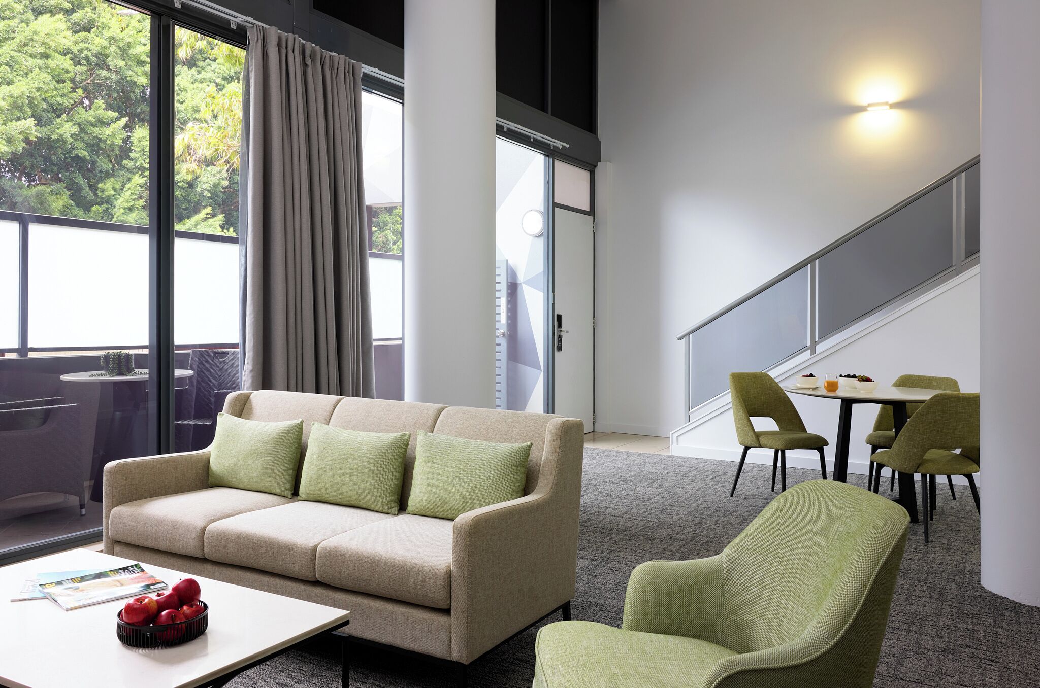 Un hotel moderno con sofá, una mesa de comedor con sillas, una mesa de centro con manzanas y ventanas amplias con vistas a árboles.