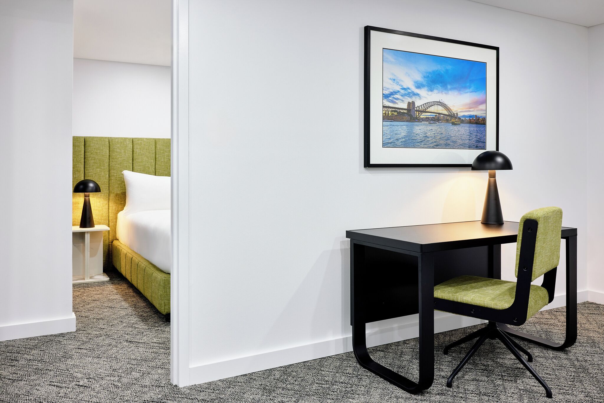 A hotel room with a bed, a desk with a chair, a framed picture on the wall, and a lamp.