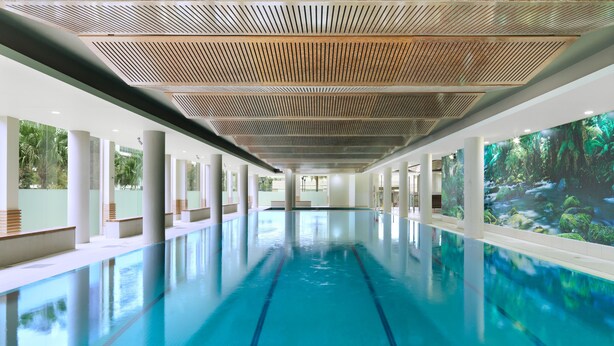Ein langer, überdachter Schwimmbereich mit klarem, blauem Wasser, umgeben von weißen Säulen und einer Holzbalkendecke.