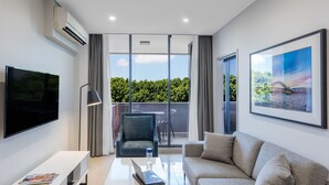 Ein modernes Wohnzimmer mit Flachbildfernseher, Sofa, Couchtisch und Sessel. Ein Balkon bietet Blick auf Grünflächen, an der Wand hängt ein gerahmtes Bild.
