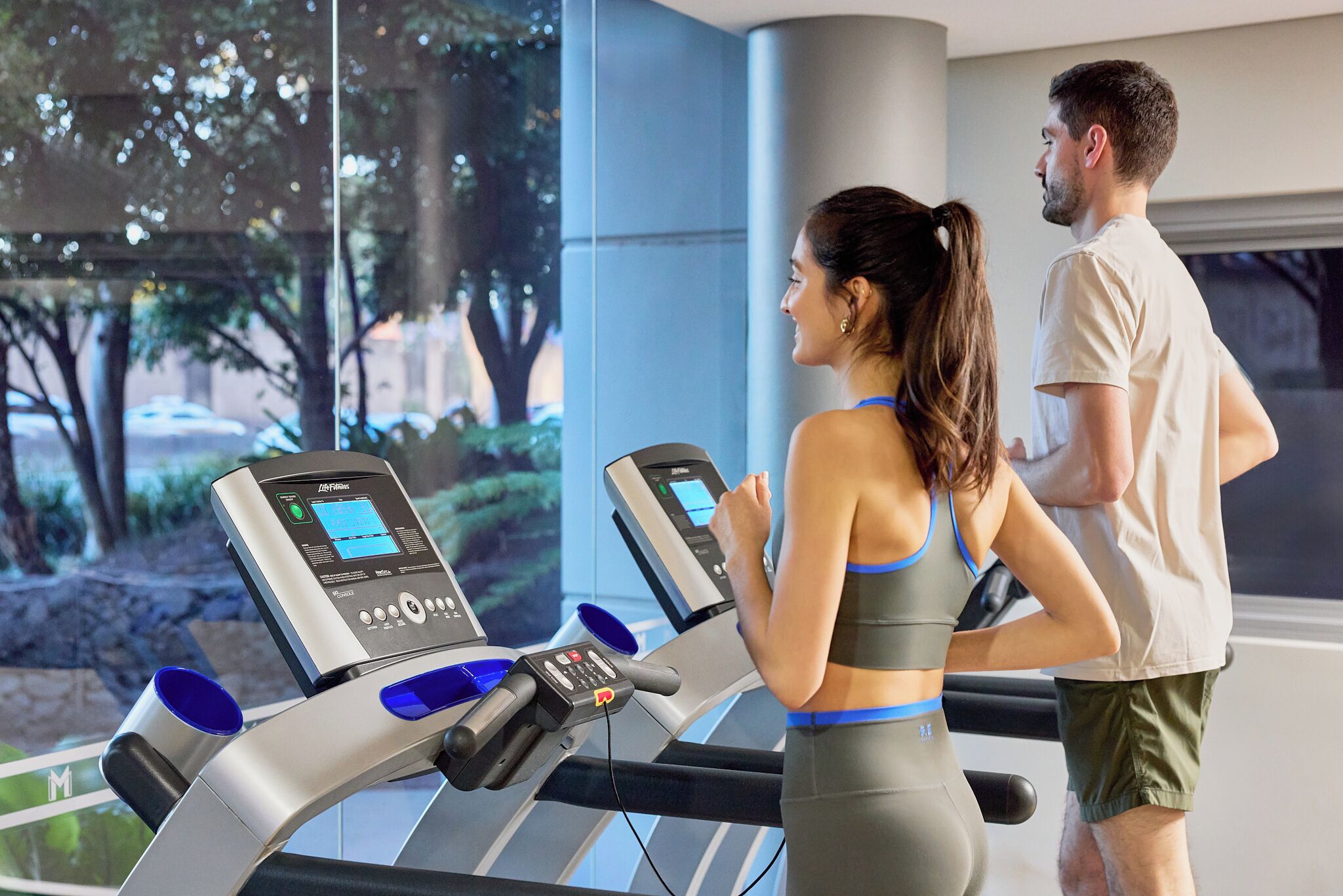 A gym with treadmills and a large window with a view of trees and a street.