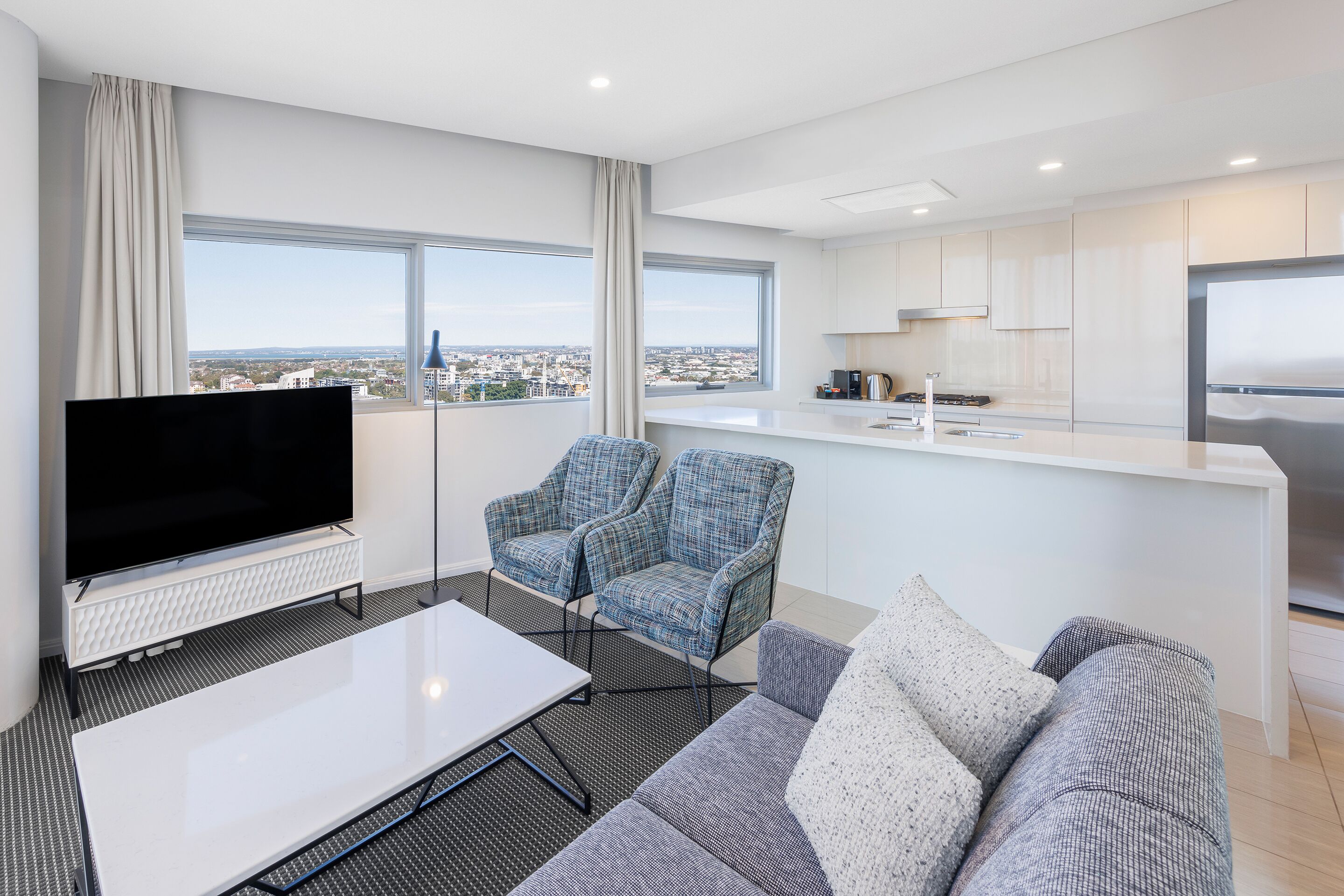 Una sala de estar moderna con una televisión de pantalla plana, un sofá, una mesa de centro y vistas de la ciudad a través de grandes ventanales.