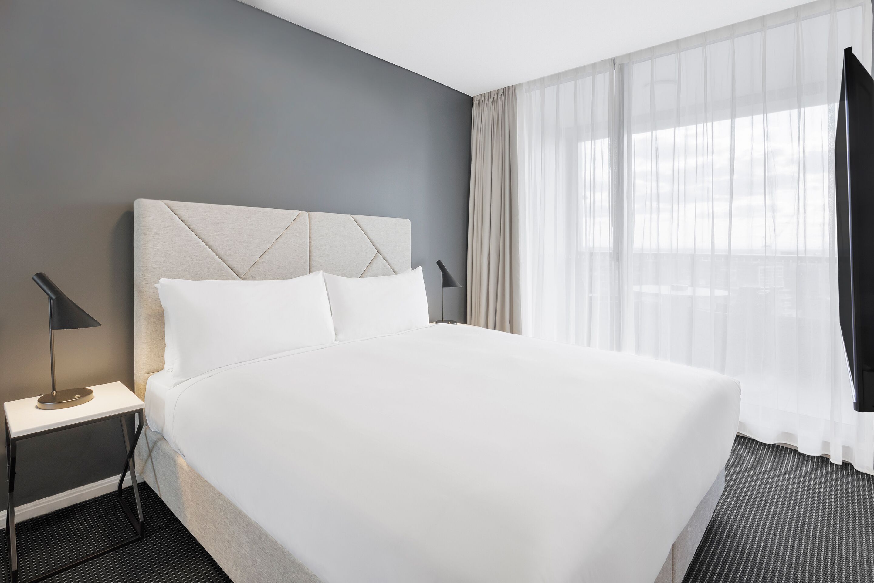 Quarto de hotel com uma cama grande, uma mesinha de cabeceira com abajur e cortinas finas na janela.