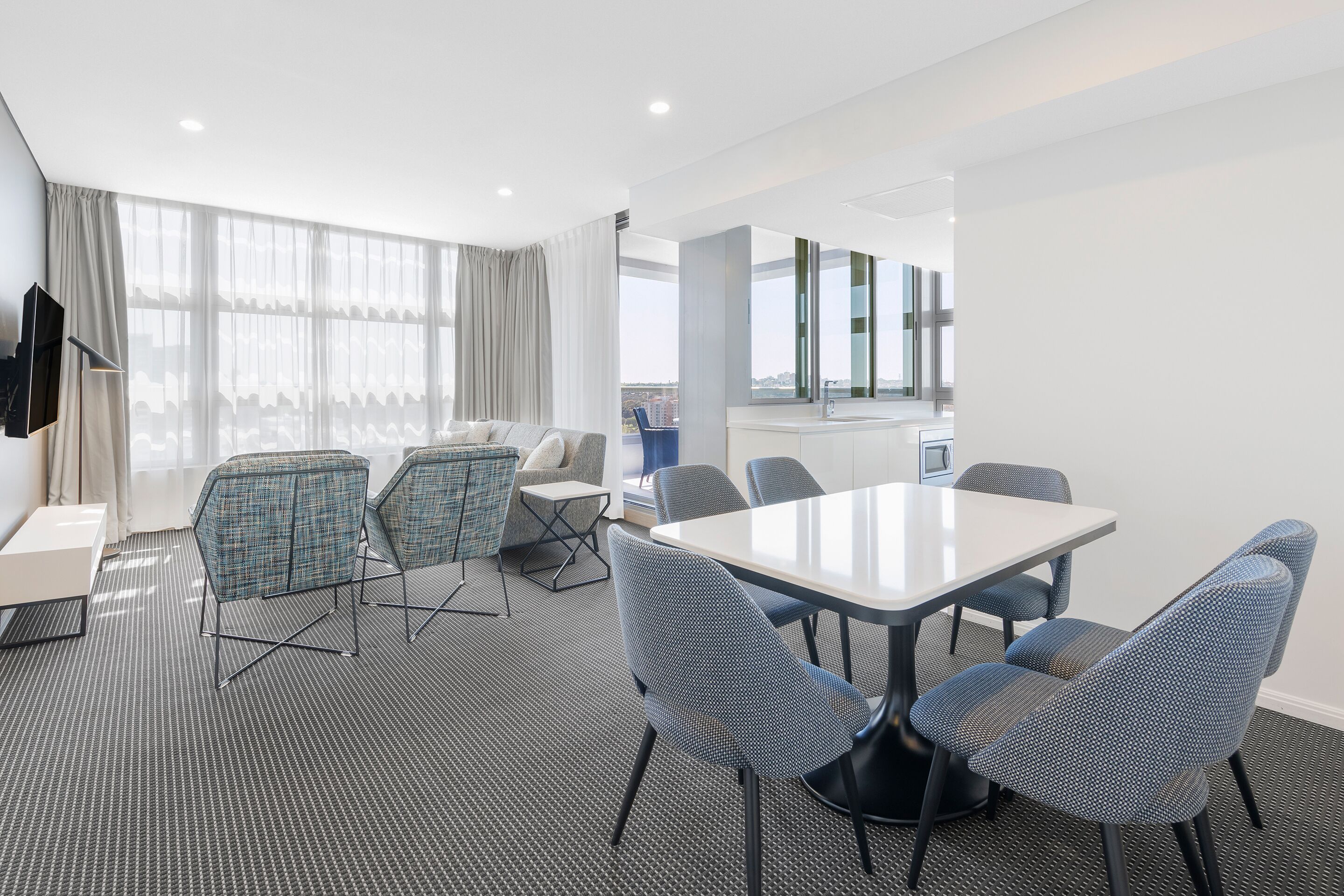 A modern hotel room with a large window, a dining area with a table and chairs, and a kitchenette.