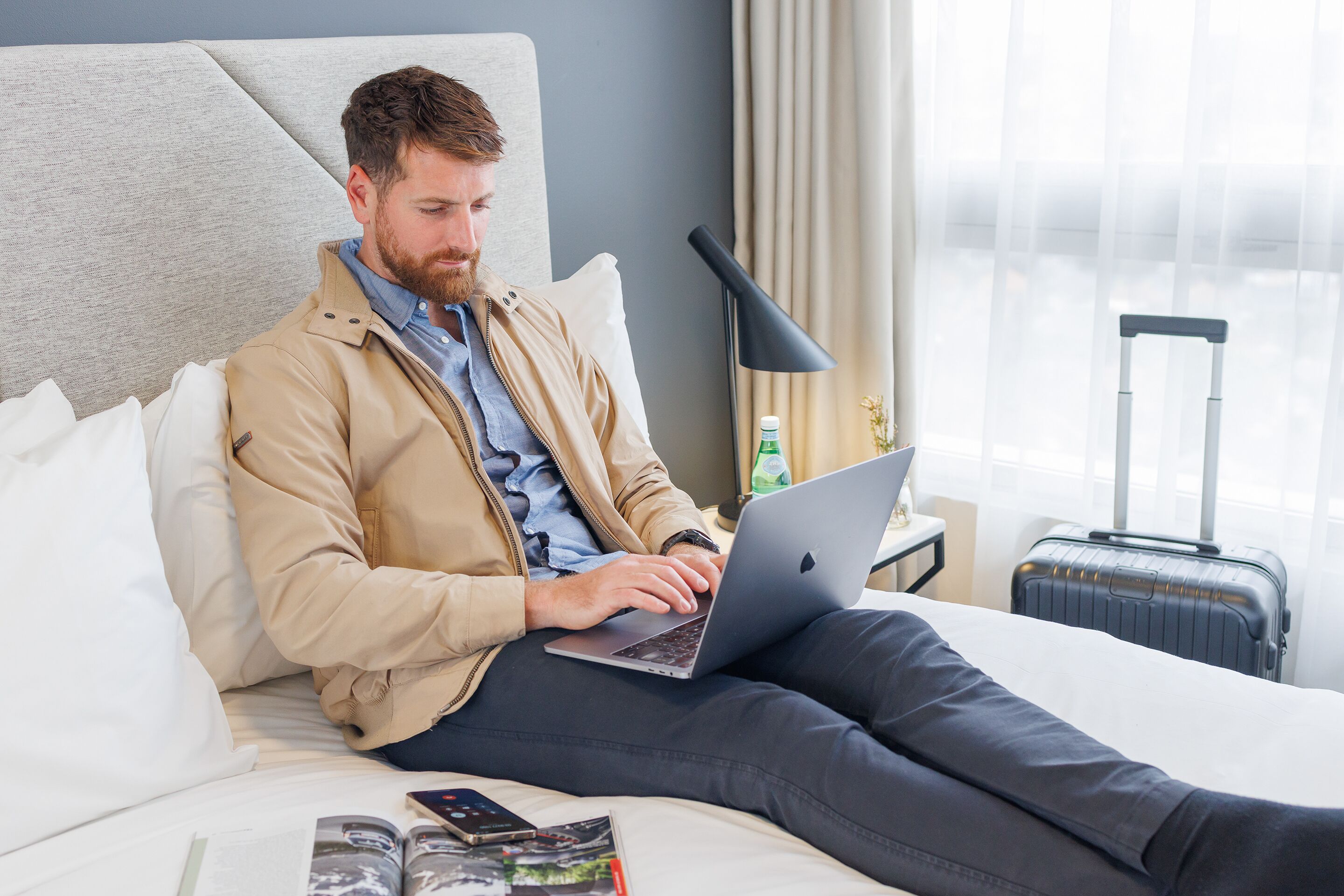 A person sitting on a bed with a laptop, a suitcase, and a bottle of water on a nightstand.