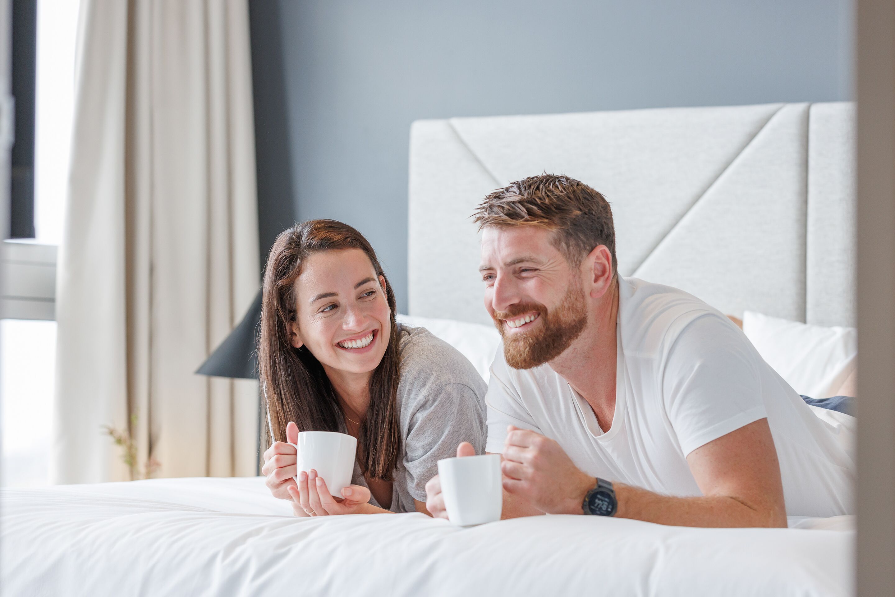 Una pareja recostada en una cama en una habitación de hotel con un cabecero moderno y una lámpara de noche.