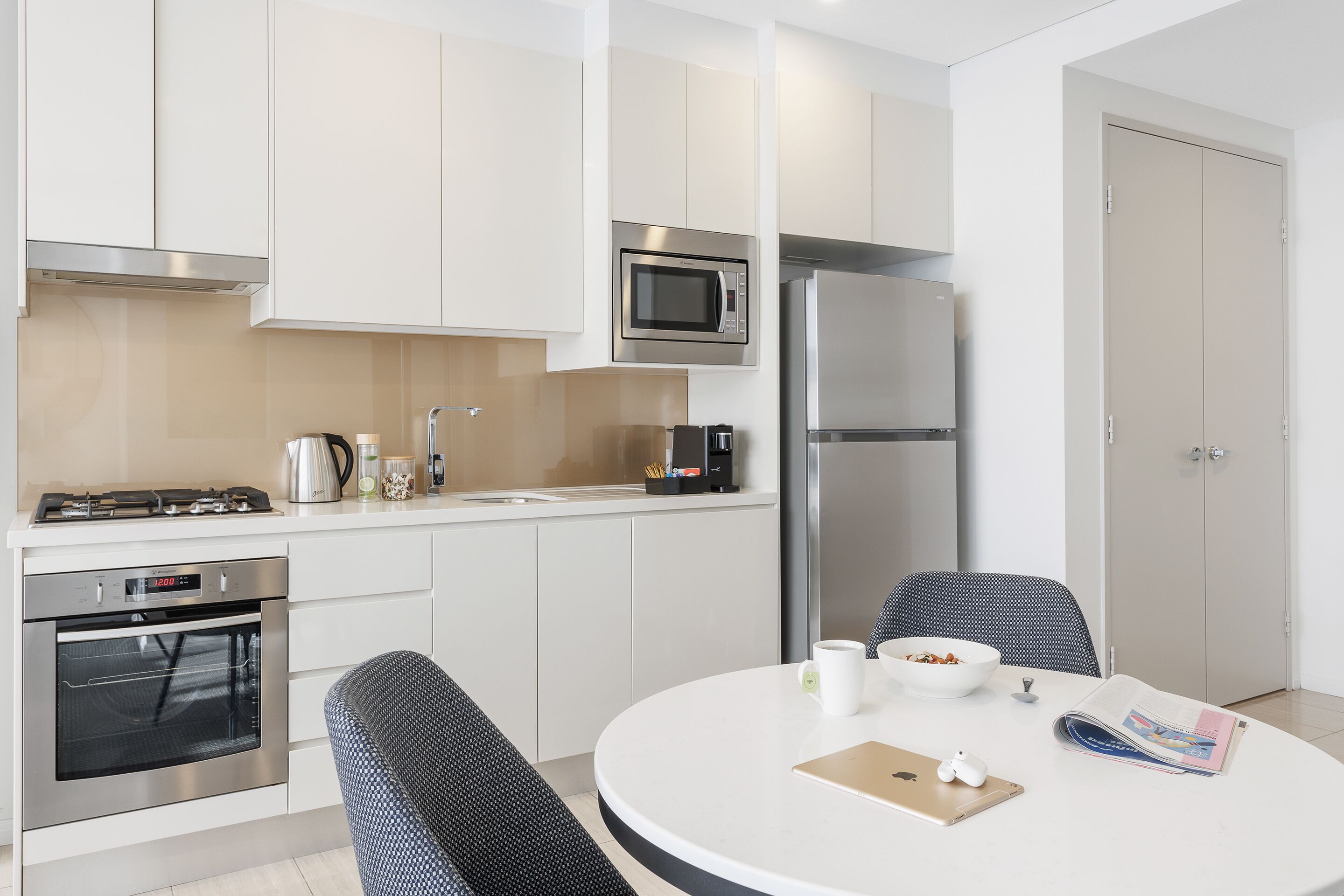 Une cuisine moderne avec des meubles blancs, un four à micro-ondes, une cuisinière et un réfrigérateur. On y trouve une table à manger avec des chaises et un saladier rempli de fruits.