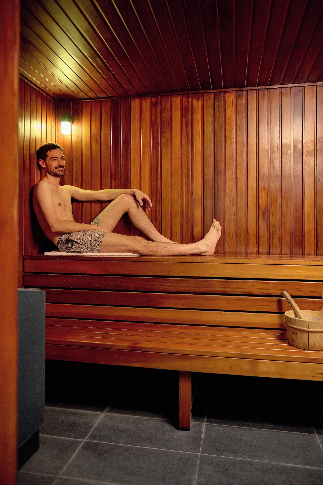 Una persona sentada en una sauna de madera con un banco y un balde de madera.