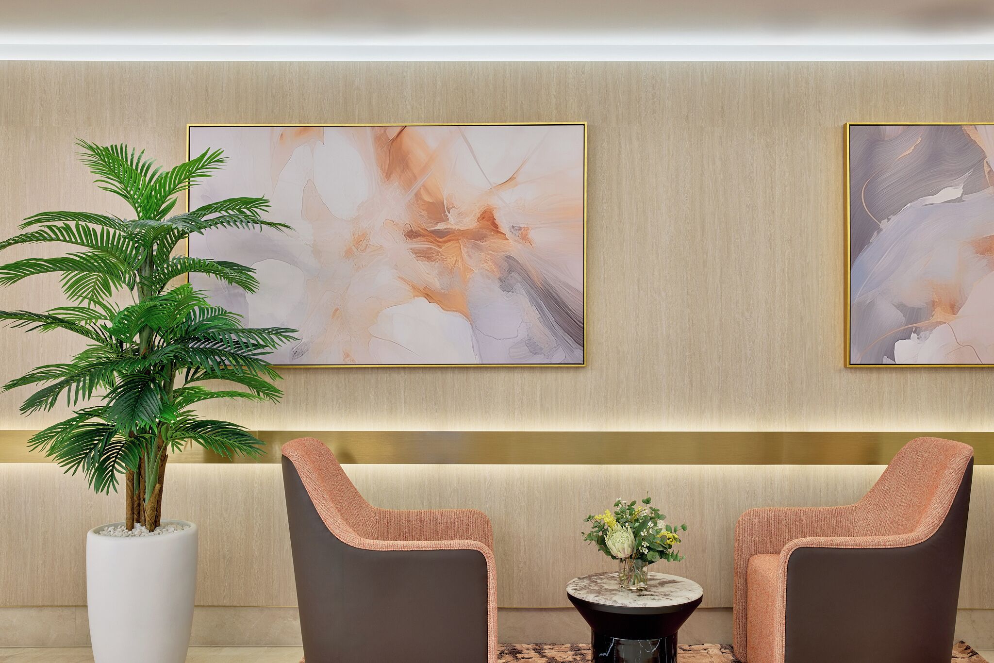A lobby with two armchairs, a potted plant, abstract paintings, and a small table with flowers.