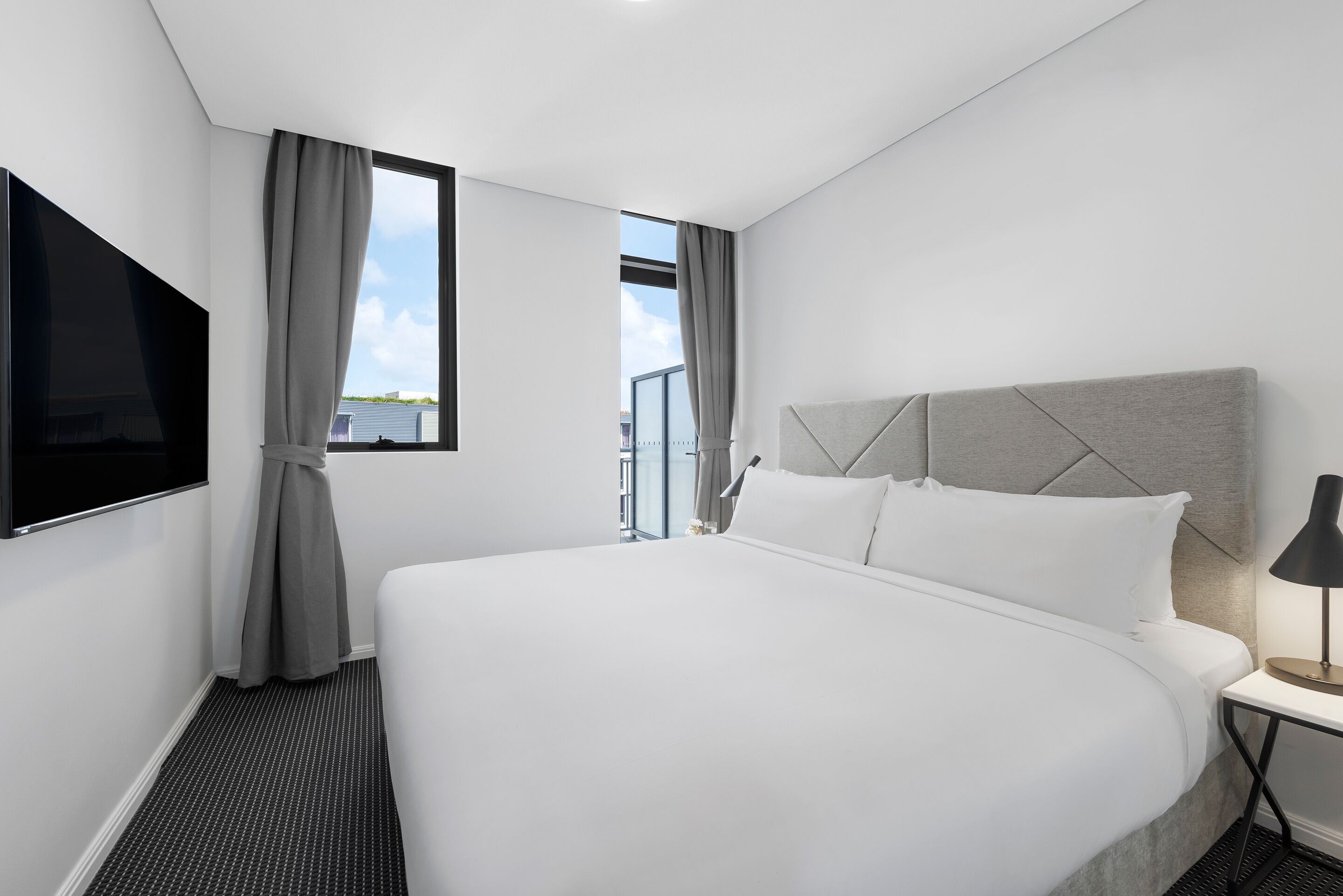 Una camera d'albergo moderna con un letto grande, una televisione a schermo piatto, un comodino con una lampada e una finestra con tende.