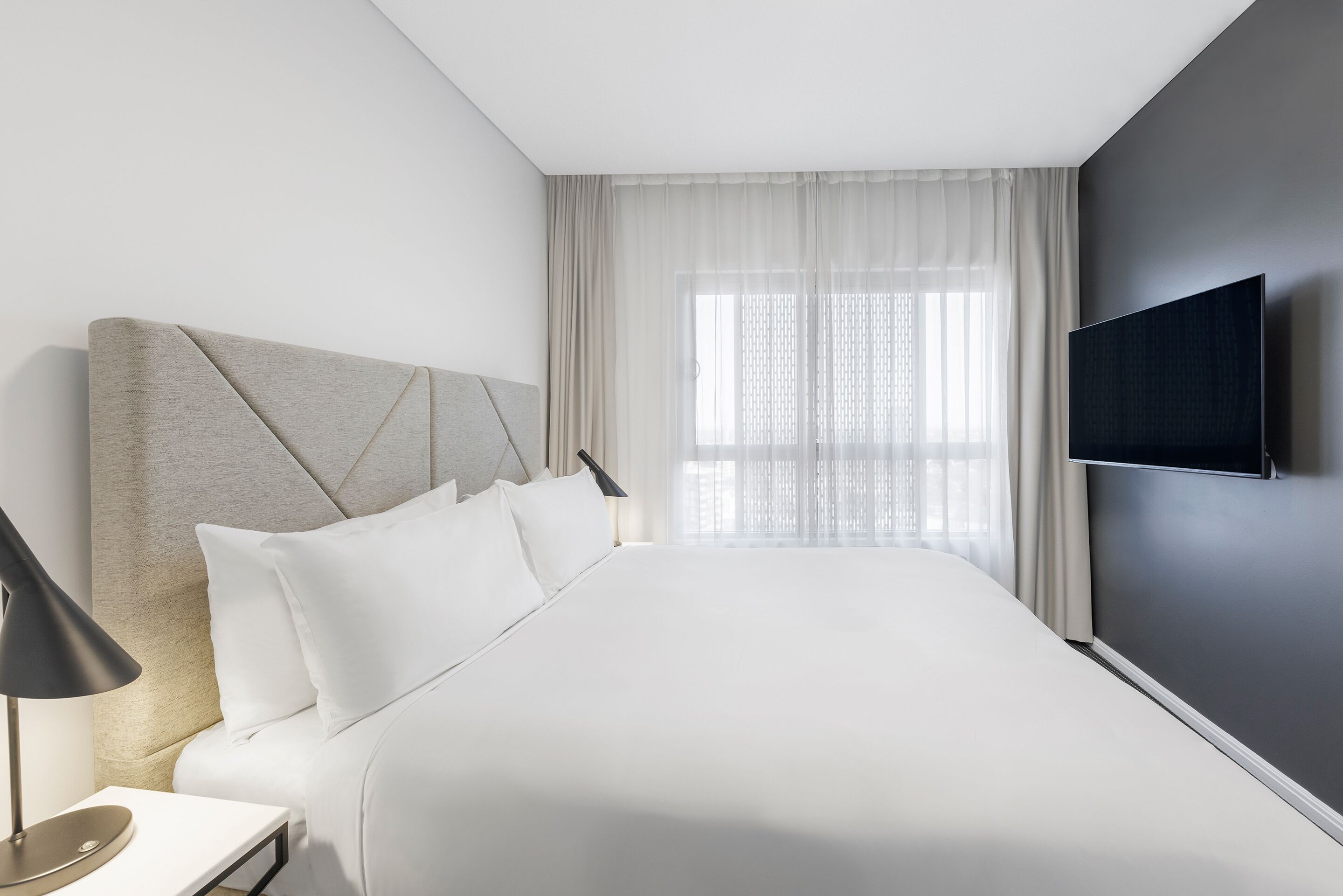 Una habitación de hotel moderna con una cama grande, un televisor de pantalla plana montado en la pared y una mesita de noche con una lámpara.