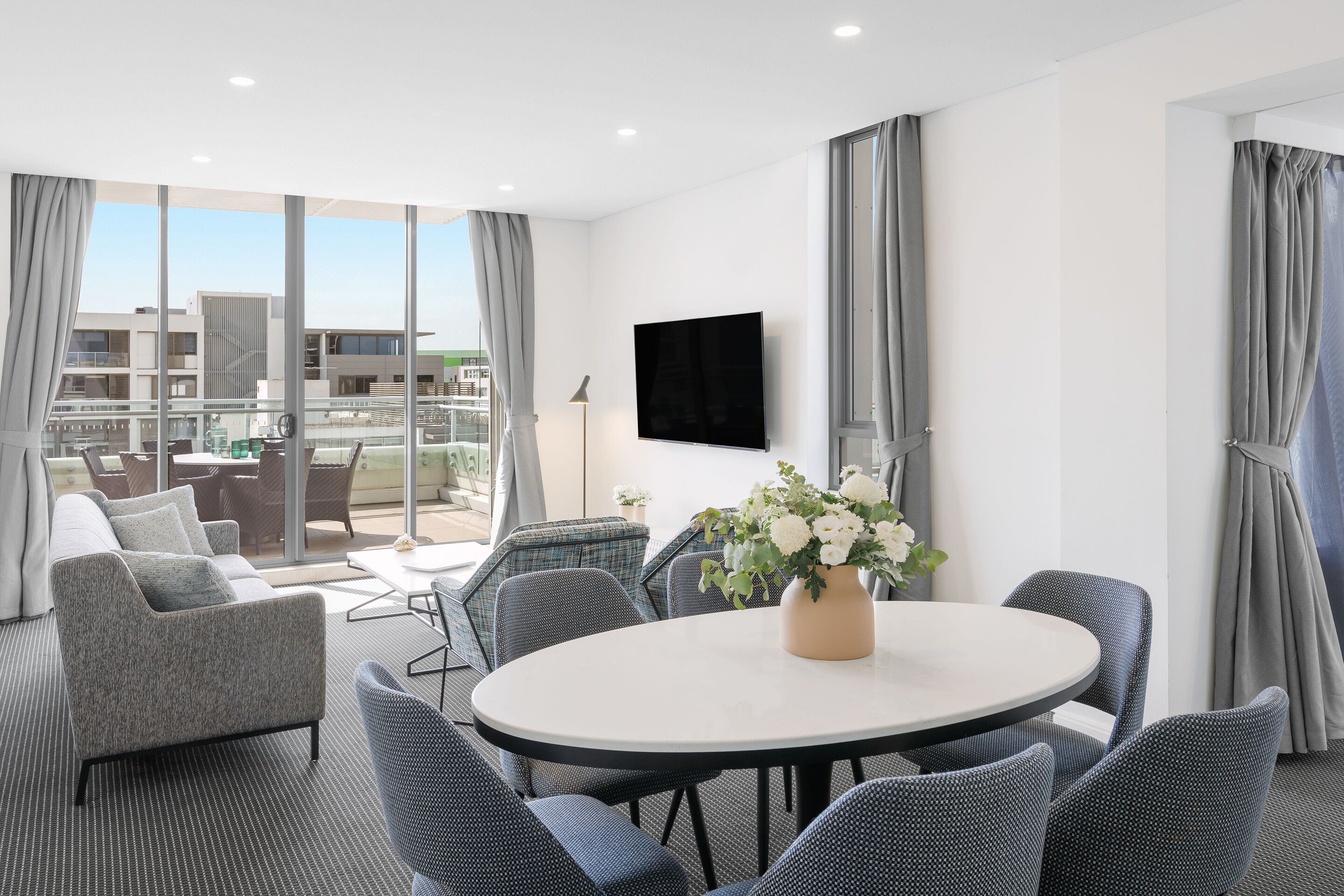 A modern hotel room with a balcony, a round table with chairs, a sofa, and a TV mounted on the wall.