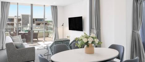 A modern hotel room with a balcony, a round table with chairs, a sofa, and a TV mounted on the wall.