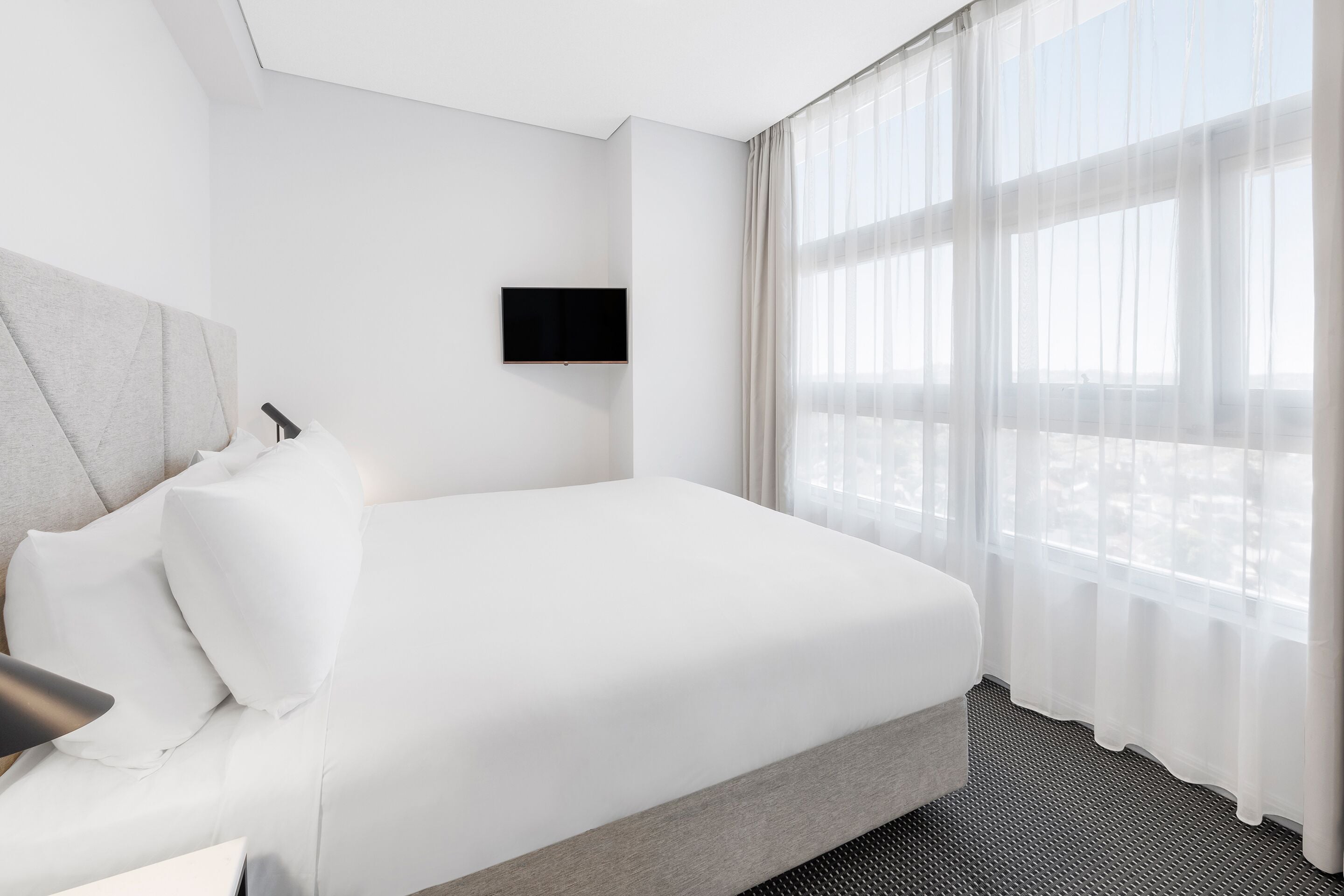 Moderni hotellihuone, jossa on suuri sänky, seinälle asennettu taulutelevisio ja suuri ikkuna, jossa on läpikuultavat verhot.