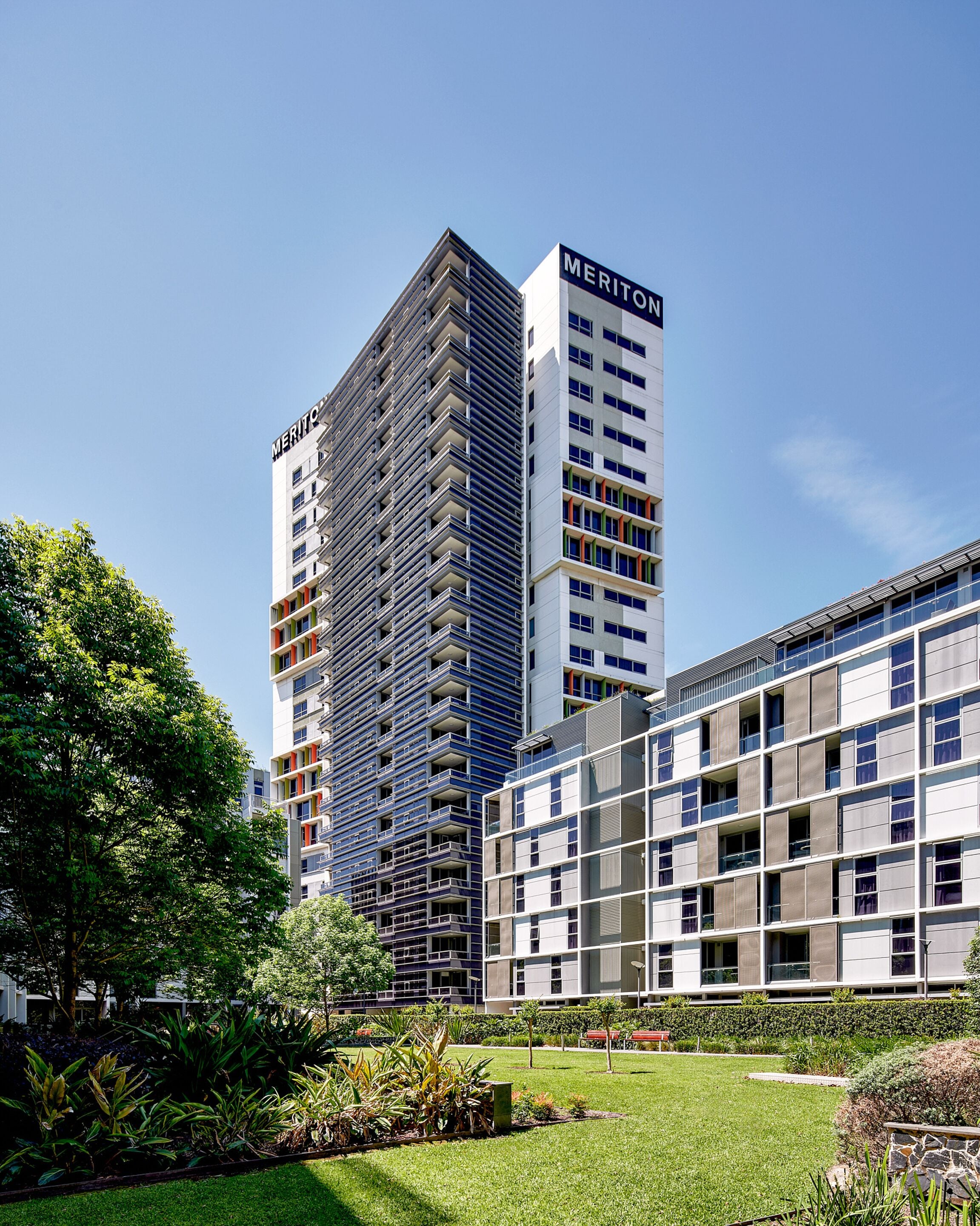Ett modernt hotell med en tydlig skylt med texten ”Meriton”, omgivet av frodig grönska och en välskött gräsmatta.