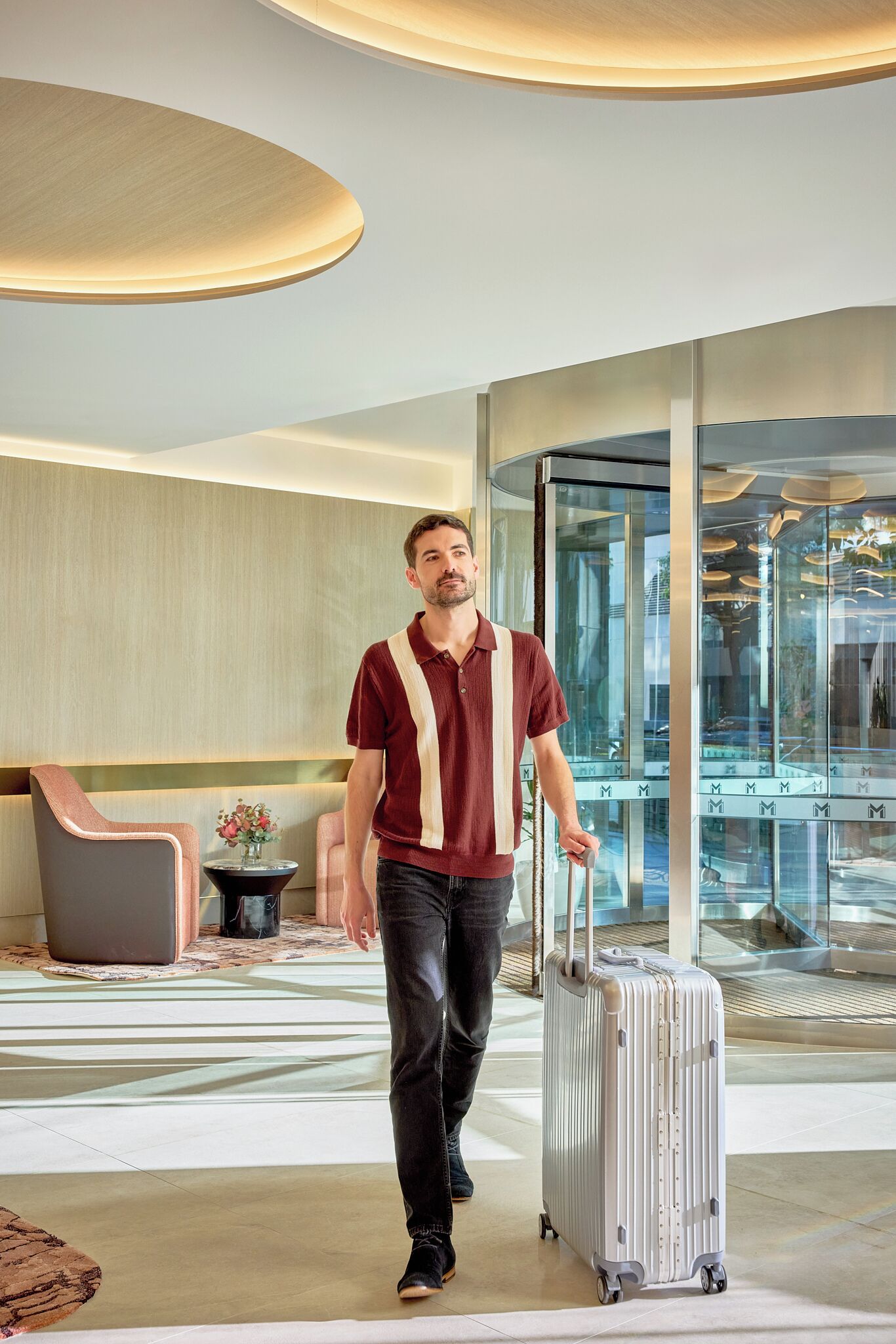 A man walking with a suitcase in a modern hotel lobby with a glass elevator and seating area.