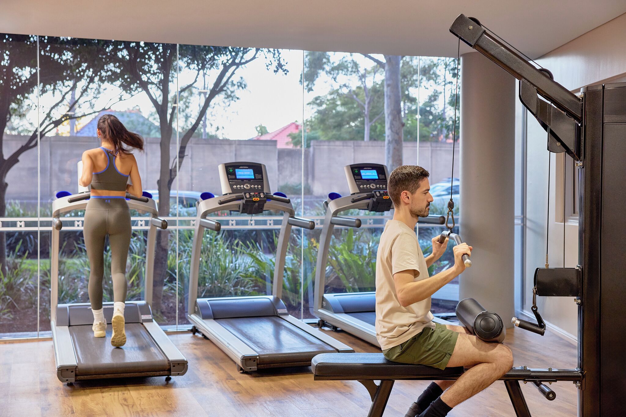 A gym with treadmills, a bench press, and a view of trees and a fence.