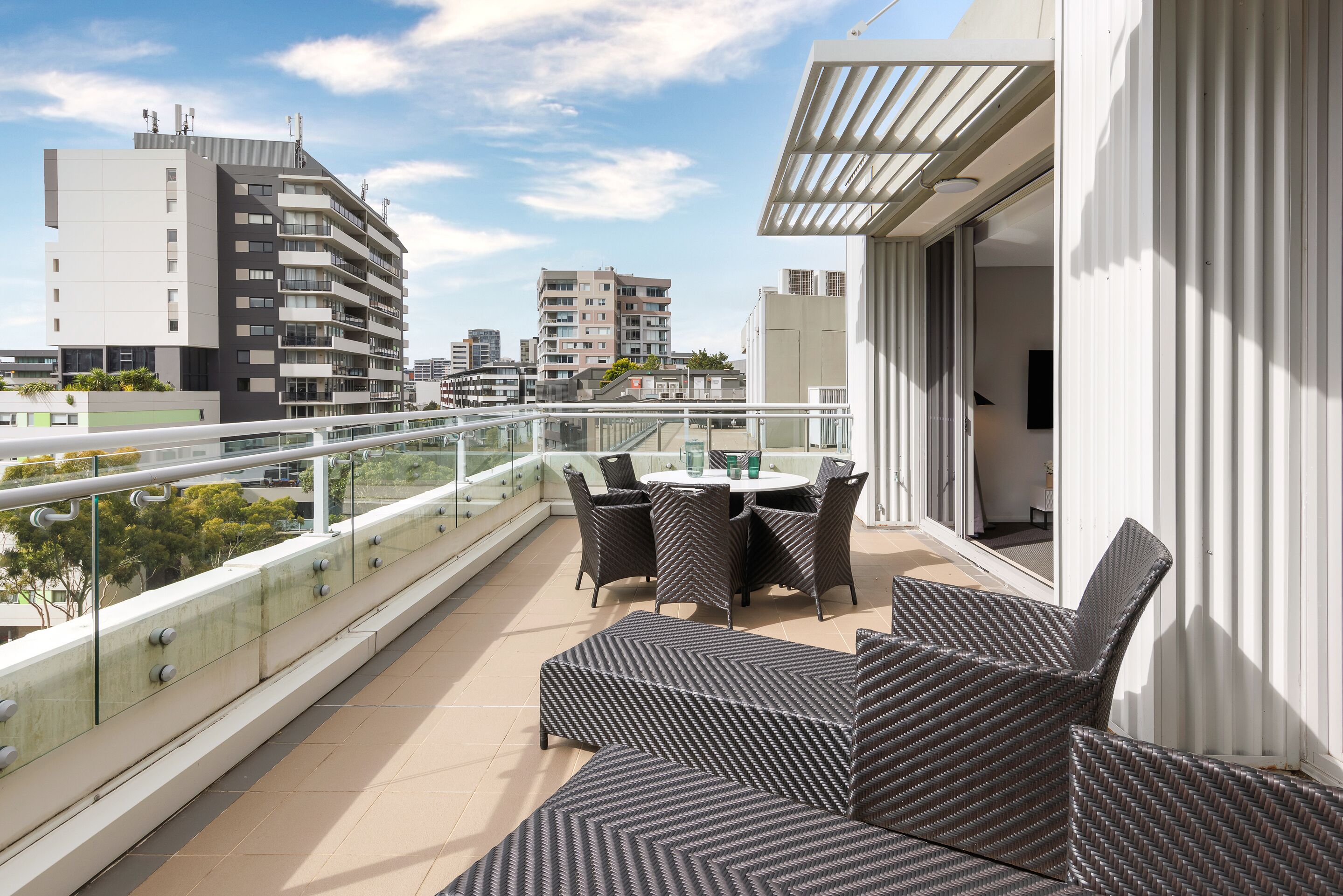 Un balcón con barandilla de vidrio, mobiliario exterior y vistas de edificios circundantes.