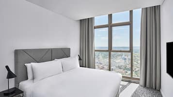 Quarto de hotel moderno com uma cama grande, uma mesa de cabeceira, uma televisão e vista para o horizonte urbano através de uma janela ampla.