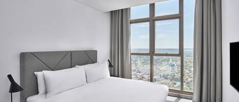 A modern hotel room with a large bed, a nightstand, a TV, and a view of a cityscape through a large window.