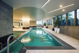 An indoor swimming pool with clear water, surrounded by a tiled floor, and a seating area with chairs and a table.
