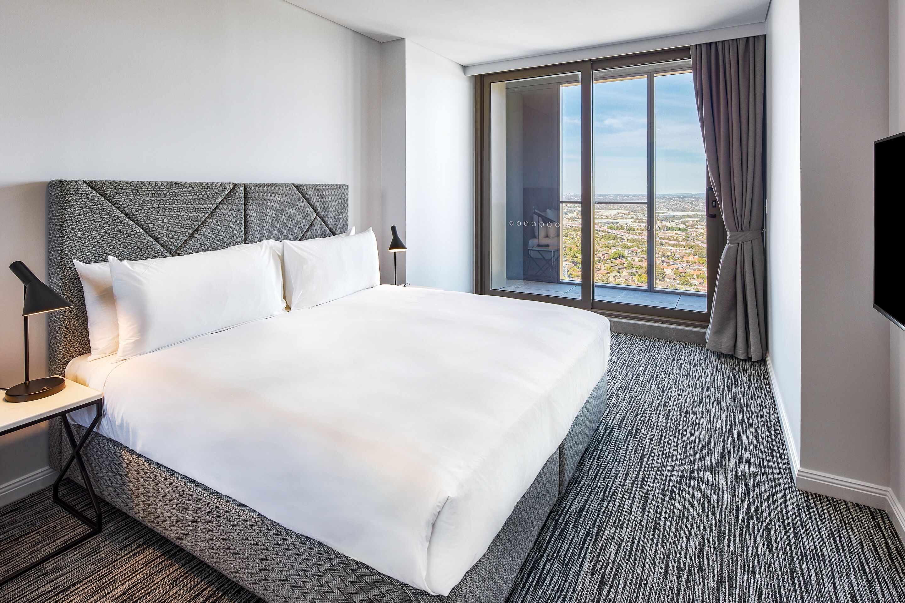 A hotel room with a large bed, a nightstand with a lamp, a view of the city through a large window, and a sliding door.