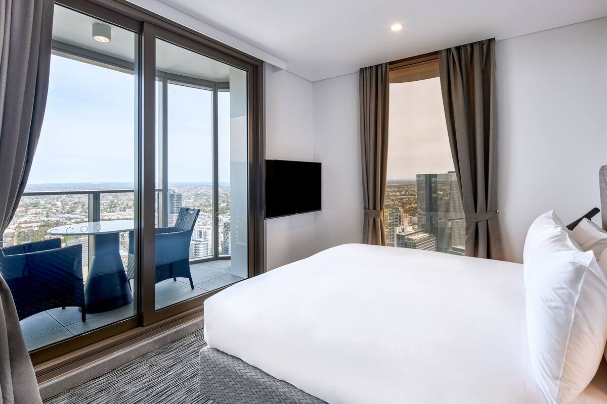 A hotel room with a large bed, a TV mounted on the wall, a balcony with a table and chairs, and a city view.
