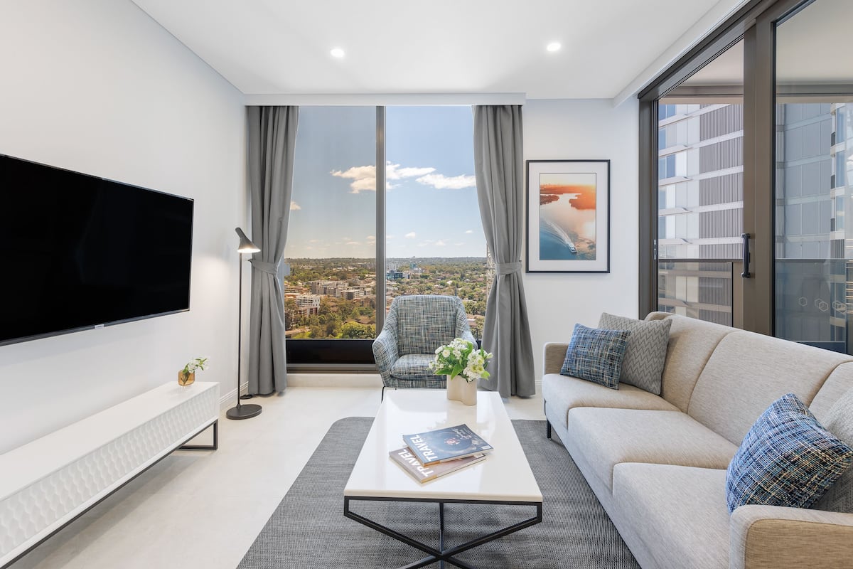 A modern living room with a large TV, a sofa, a coffee table, and a view of the city through large windows.