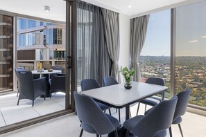 A modern dining area with a round table and six chairs, a large window with a city view, and a balcony with a dining set. - Meriton Suites George Street, Parramatta (Parramatta)