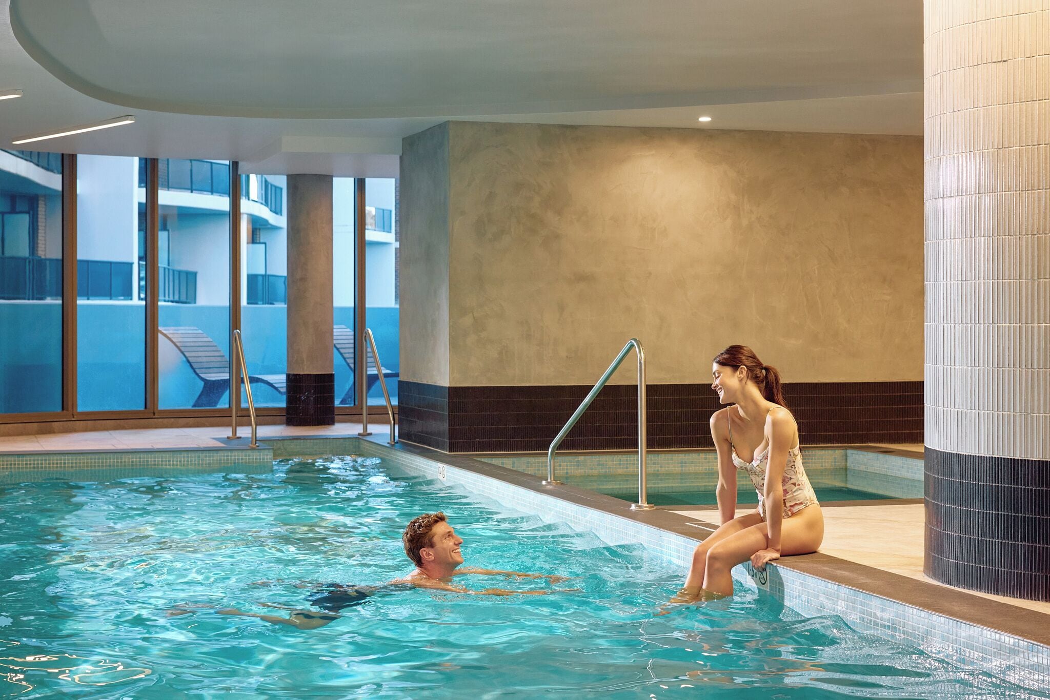 Una piscina interior con agua cristalina, un hombre y una mujer en la piscina, y vistas de un edificio exterior.