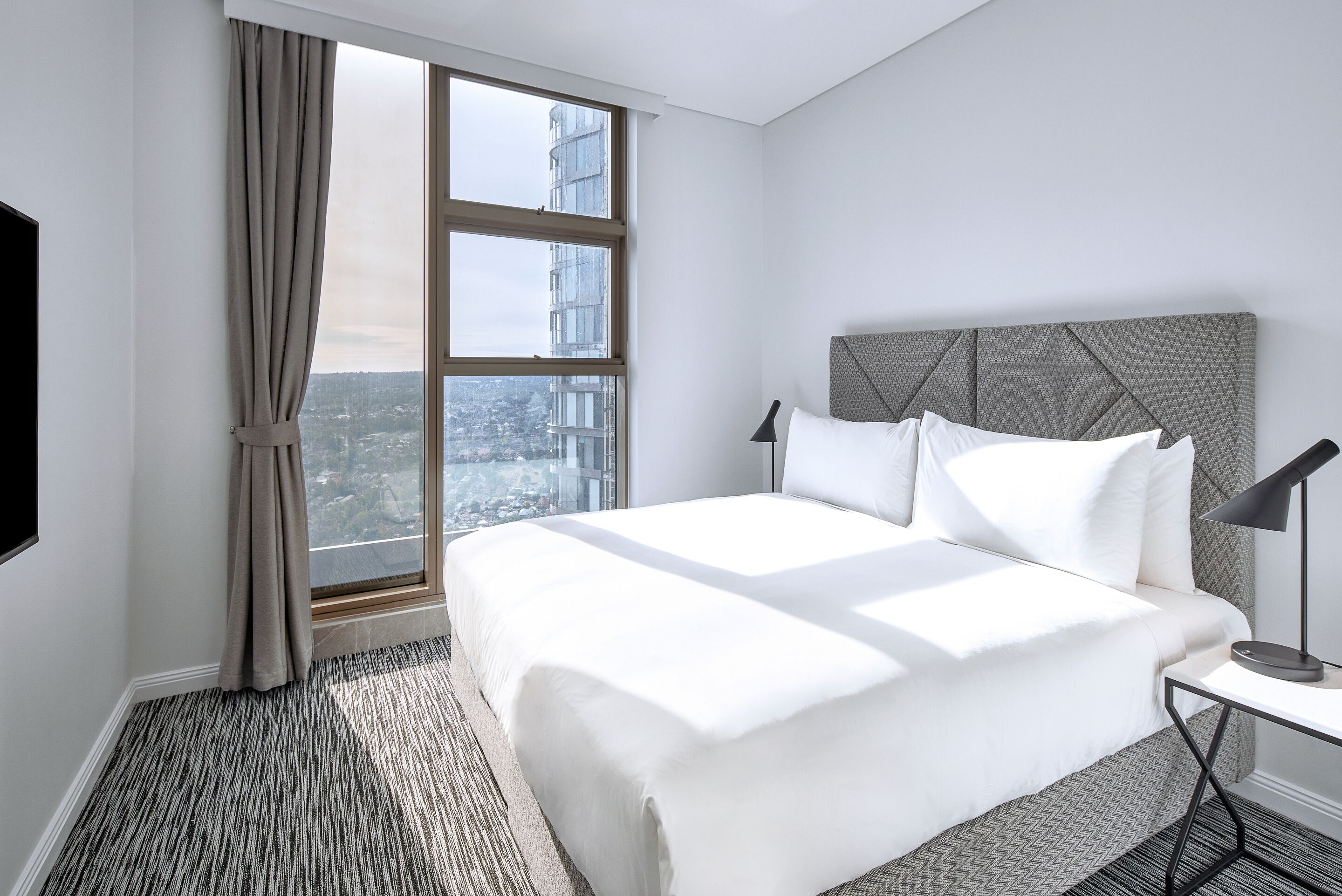 A hotel room with a large bed, a nightstand, a lamp, and a view of a cityscape through a large window.