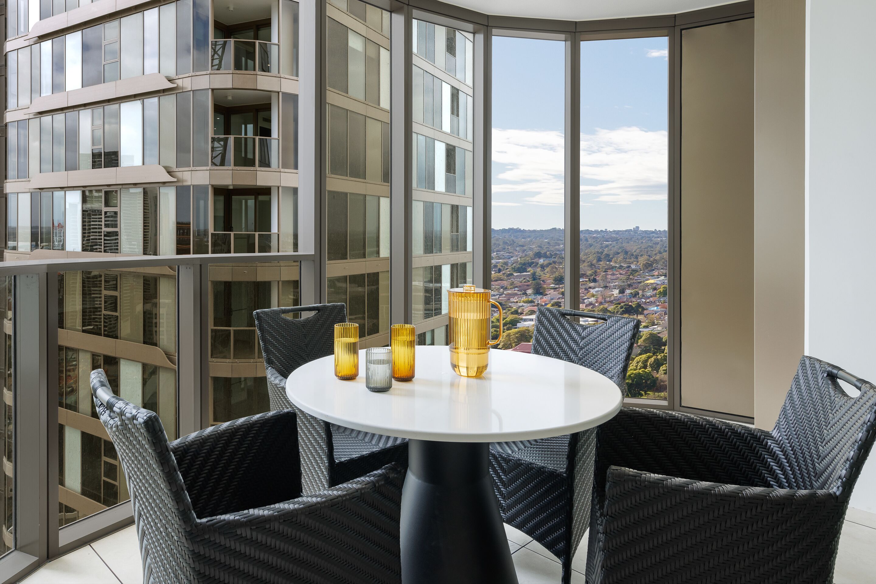 Ein Balkon mit rundem Tisch und Korbstühlen, mit Blick auf eine Stadtlandschaft.