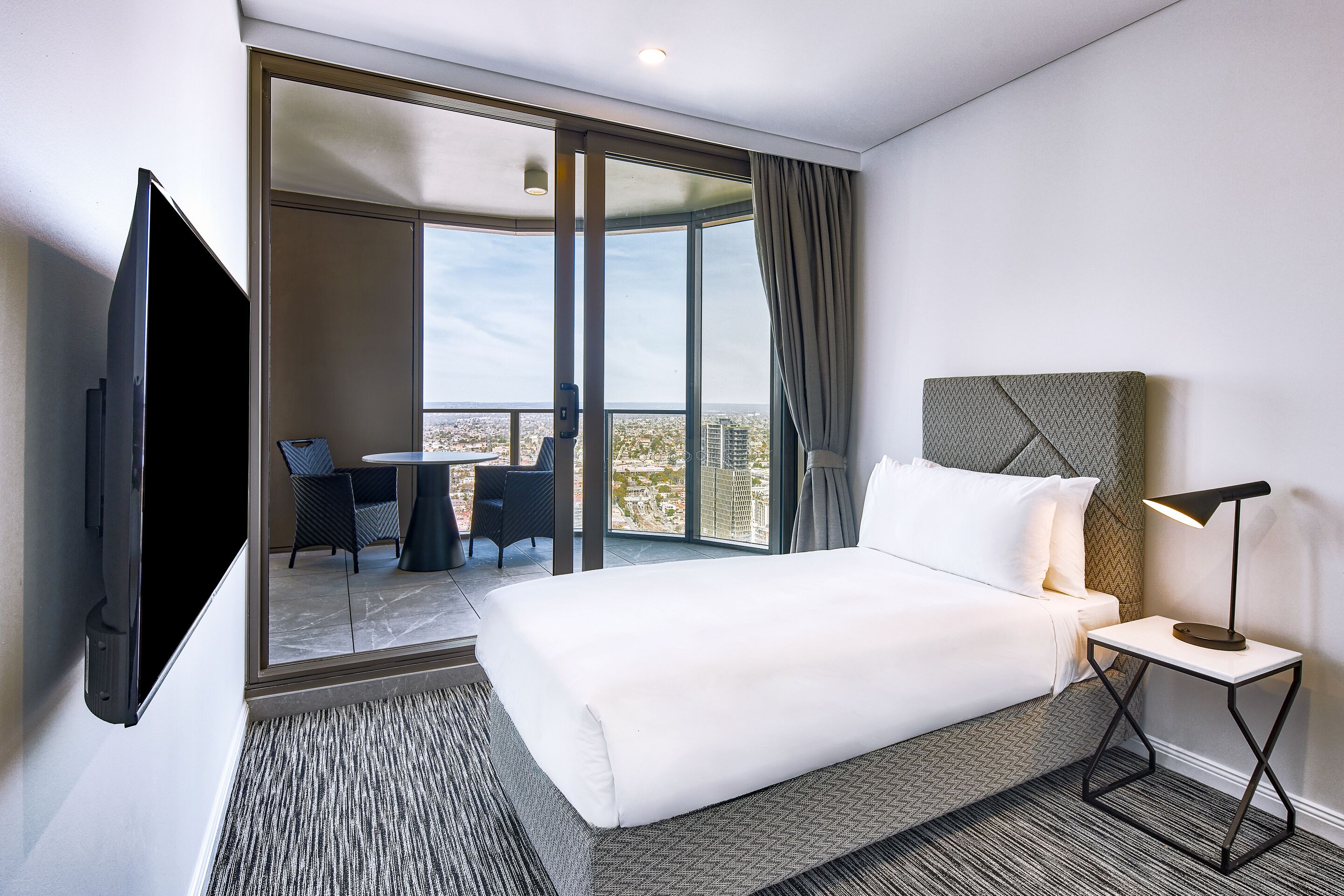 A hotel room with a large bed, a TV, a desk with a chair, and a balcony with a view of the city.