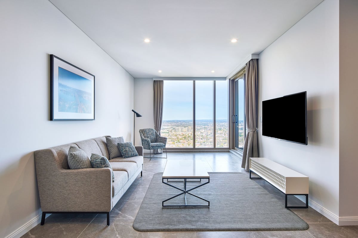 A modern living room with a sofa, a coffee table, a TV, and a large window offering a city view.