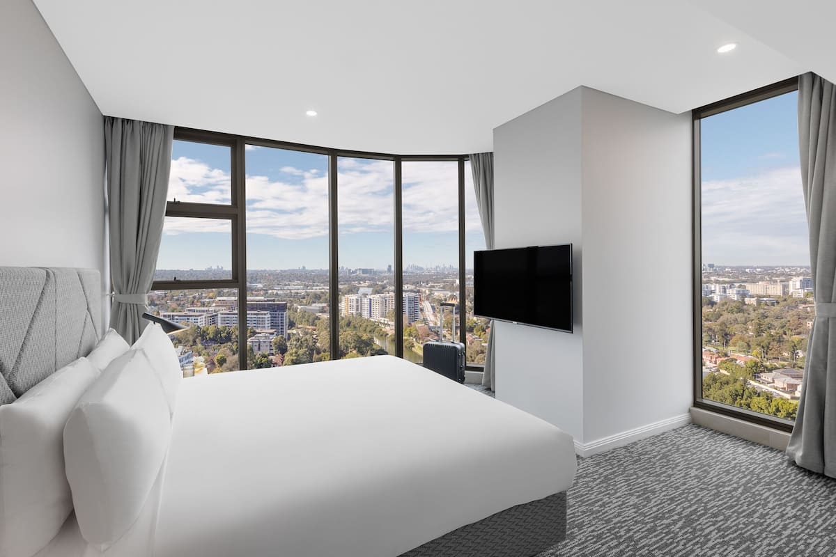 A modern hotel room with a large bed, a flat-screen TV, and a city view through large windows.