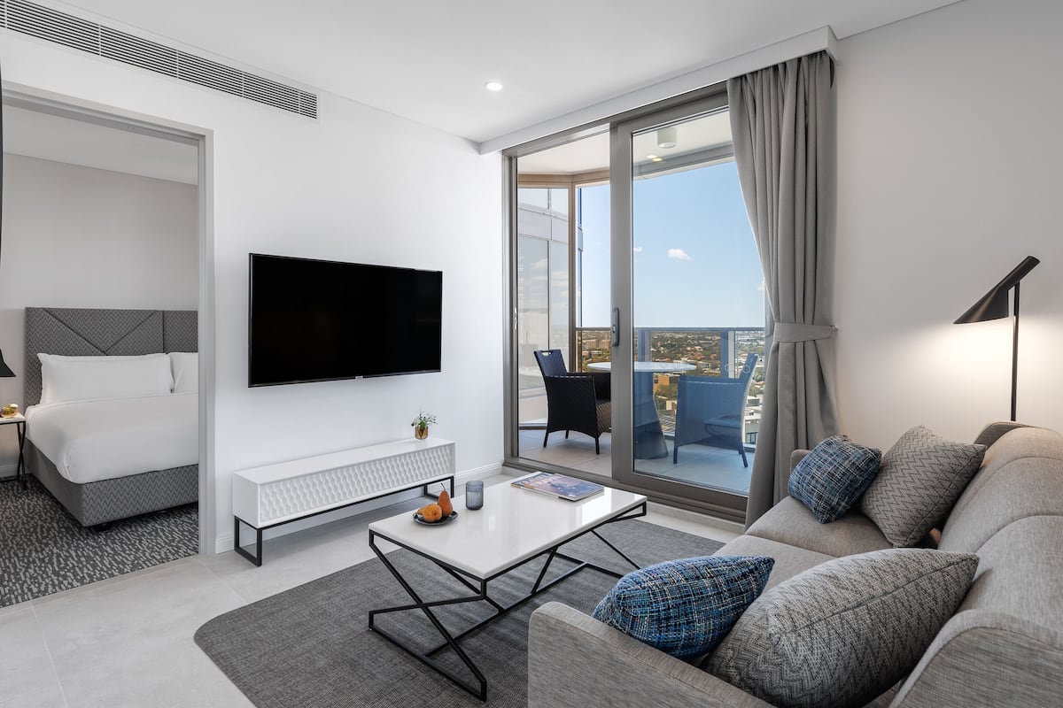 A modern living room with a flat-screen TV, a sofa with patterned cushions, a coffee table, and a view of the city through large glass doors.