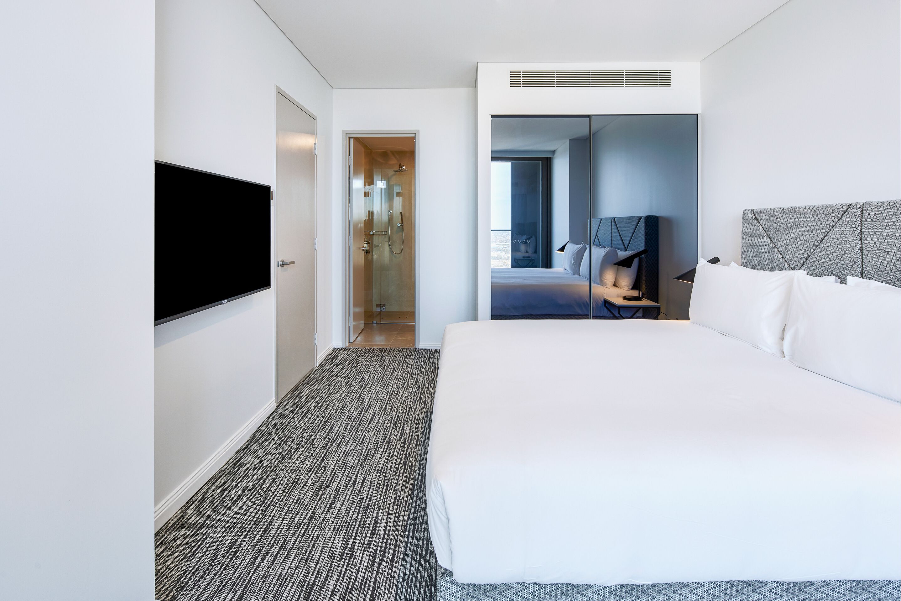 Ett modernt hotellrum med en stor säng, en platt-TV som är monterad på väggen och en duschavdelning som syns genom en glasdörr.
