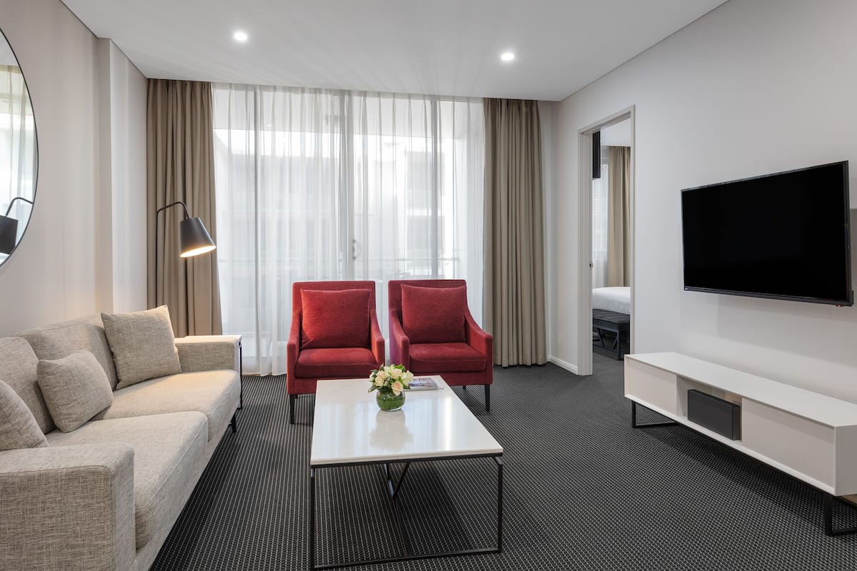 A modern hotel room with a beige sofa, two red armchairs, a white coffee table with a vase, a flat-screen TV, and a large window with sheer curtains.