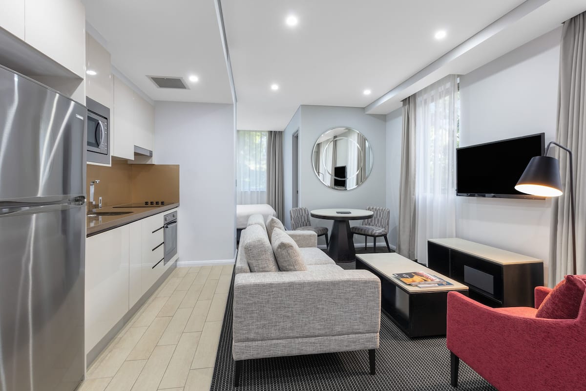 A modern hotel room with a kitchenette, a sofa, a dining table, a TV, and a mirror.