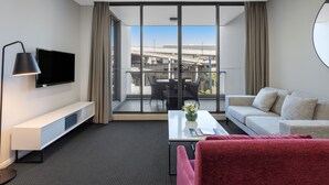 Una habitación de hotel moderna con un balcón amplio, un televisor de pantalla plana, un sofá, un sillón rojo y una mesa de centro con un florero.