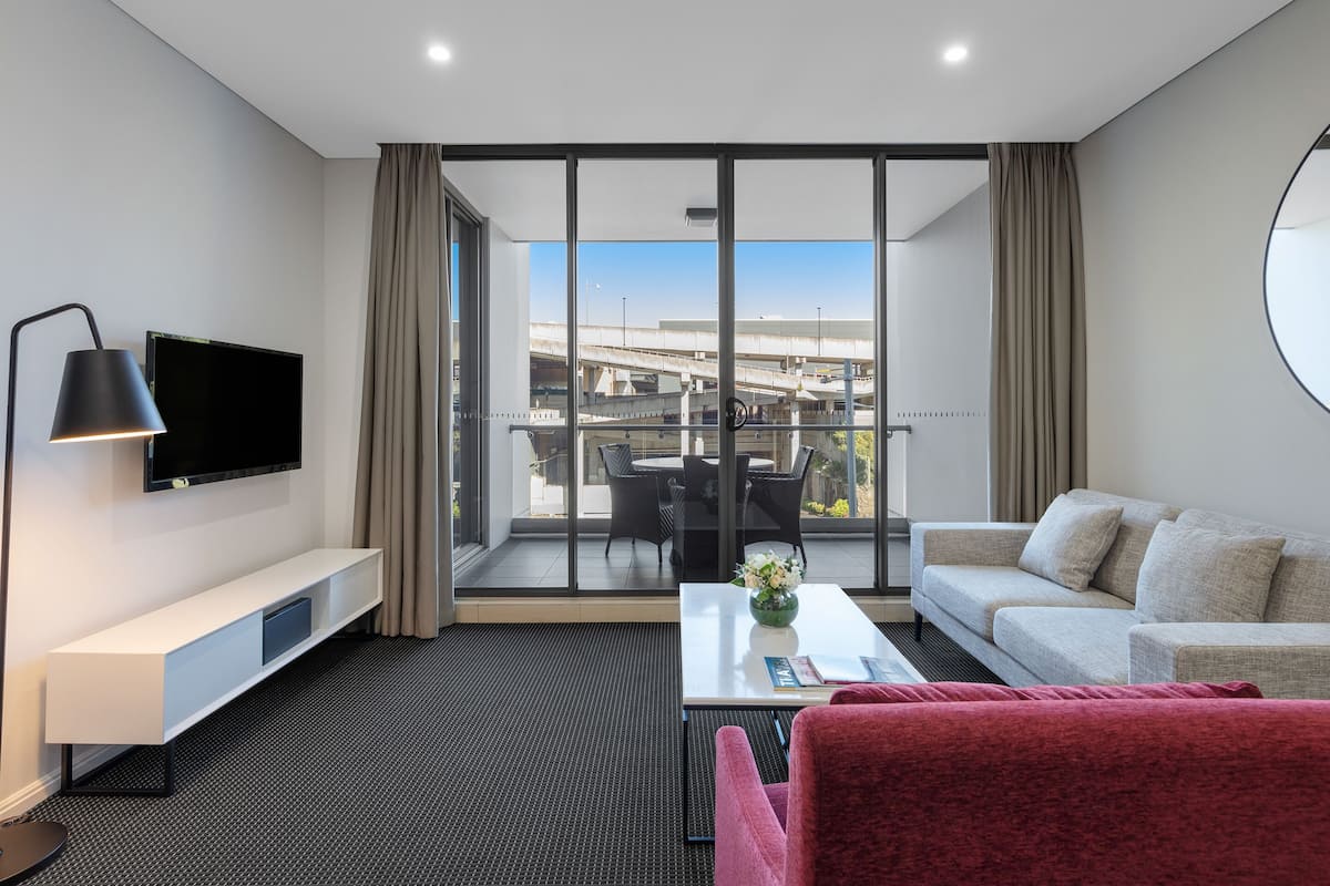 A modern hotel room with a large balcony, a flat-screen TV, a sofa, a red armchair, and a coffee table with a vase.
