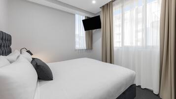 A hotel room with a large bed, a flat-screen TV mounted on the wall, and sheer curtains on the windows.