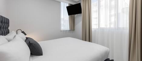 A hotel room with a large bed, a flat-screen TV mounted on the wall, and sheer curtains on the windows.